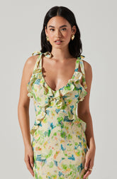 A woman wearing a sleeveless dress with shoulder ties. The dress features a colorful, abstract floral pattern with shades of green, blue, yellow, and pink. The neckline is decorated with ruffles that extend down the front of the dress. She is accessorized with large, circular gold earrings.